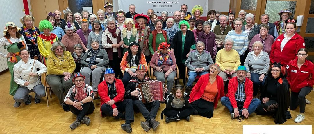 Eine Gruppe von Menschen in farbenfrohen Kostümen und Hüten posiert für ein Foto bei einer Veranstaltung des Roten Kreuzes in Waidhofen. Einige sitzen auf Stühlen, während andere stehen und alle lächeln in die Kamera.
