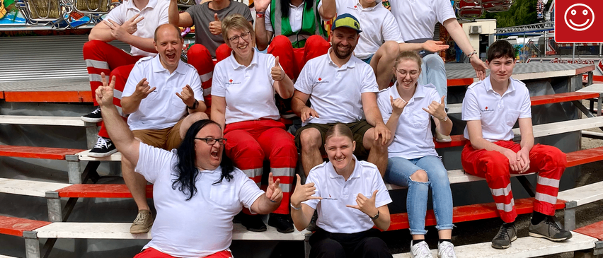 Eine Gruppe von Freiwilligen, gekleidet in weißen und roten Sicherheitsuniformen, posiert für ein Foto auf einer Tribüne. Einige machen Friedenszeichen. Ein Karussell ist im Hintergrund zu sehen.