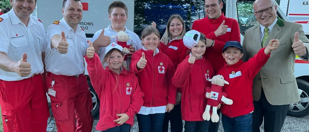 A group of smiling children and adults, all dressed in red jackets, posing for a photo with an ambulance in the background.