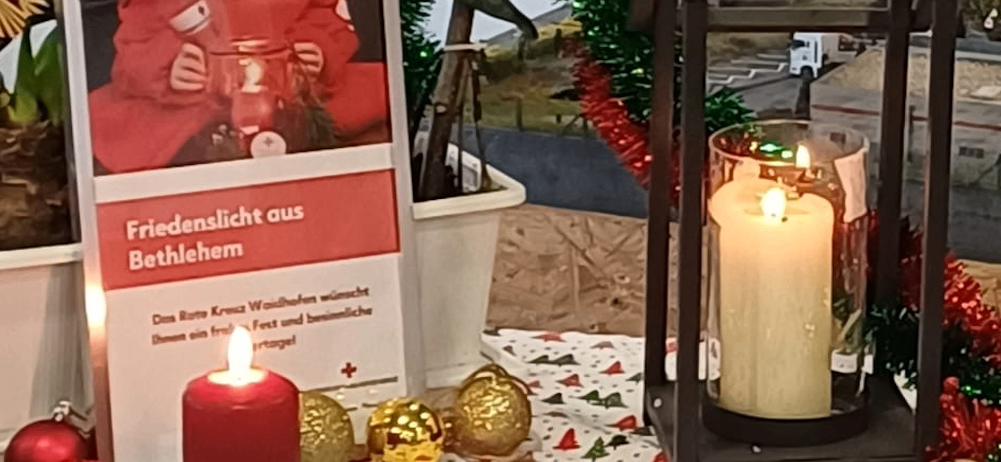 Eine Weihnachtsdekoration mit einer roten Kerze, einem Schild, das Friedenslicht aus Bethlehem steht, und einem Laternen mit brennender Kerze.