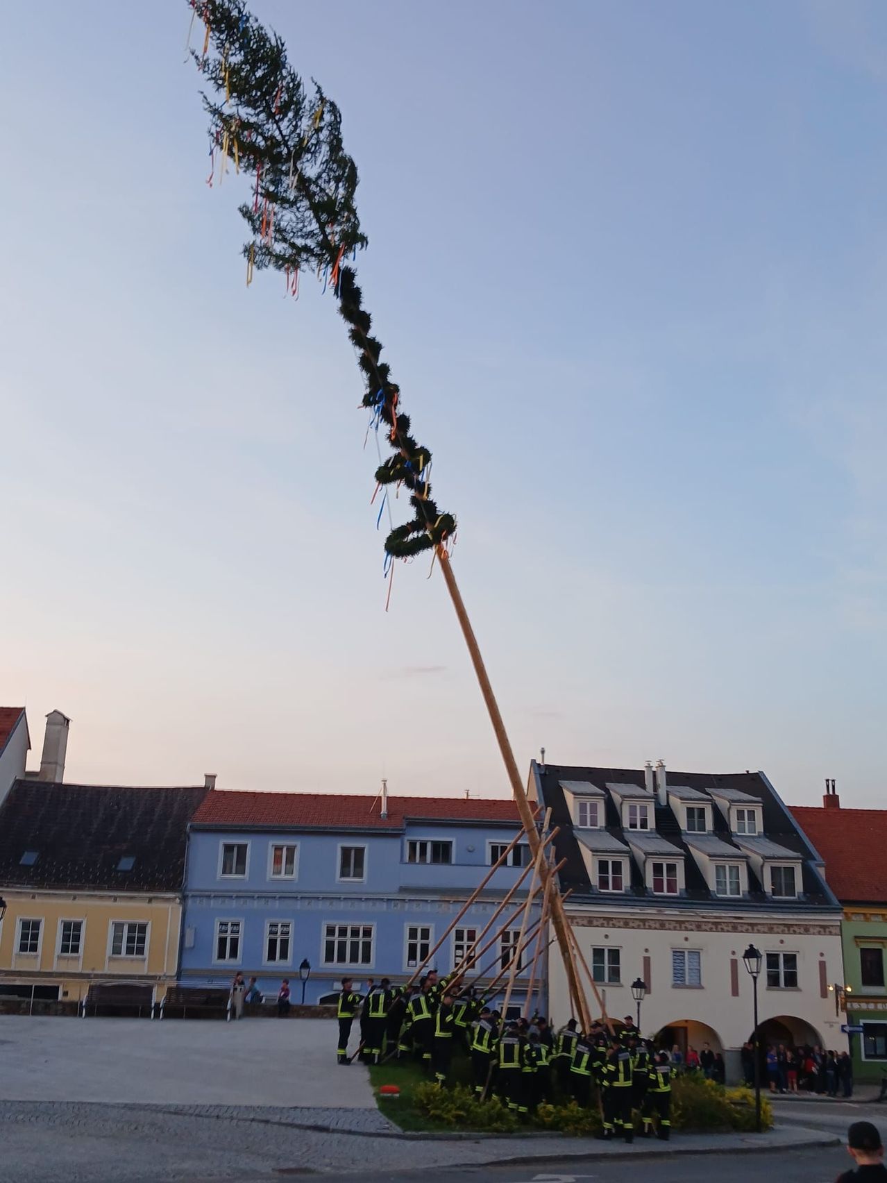 Ein hoher Baumzweig, geschmückt mit Bändern und Ornamenten, wird von einer Gruppe von Menschen in Sicherheitswesten in einem Stadtplatz mit Gebäuden im Hintergrund angehoben.