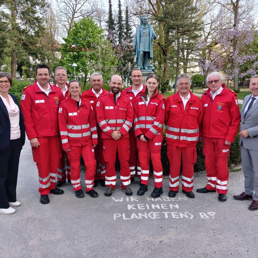 Eine Gruppe von Menschen in roten Uniformen mit reflektierenden Streifen steht vor einer Statue. Sie posieren für ein Foto mit Bäumen und Büschen im Hintergrund.