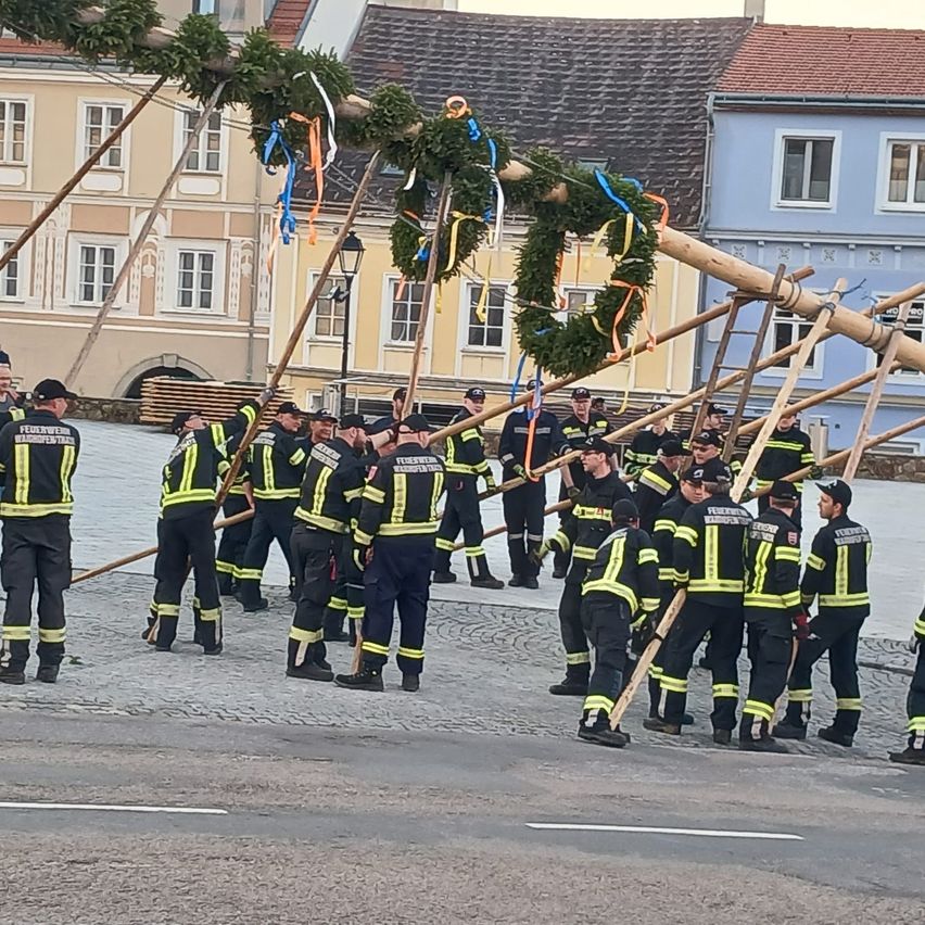 Eine Gruppe Feuerwehrleute in Uniform steht auf einer Kopfsteinpflasterstraße mit Holzstangen und Girlanden. Im Hintergrund befinden sich Gebäude mit verschiedenen Fenstern.