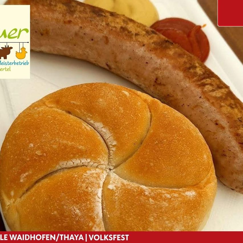 Eine Wurst und ein rundes Brot auf einem weißen Teller. Die Wurst hat eine goldbraune Kruste und wird von einer Scheibe roter Zwiebel begleitet. Das Brot hat eine goldene Farbe und ist in vier Abschnitte geschnitten. Das Bild enthält auch einen Wasserzeichen mit dem Text 'Le Waidhofen/Thaya | Volksfest'.