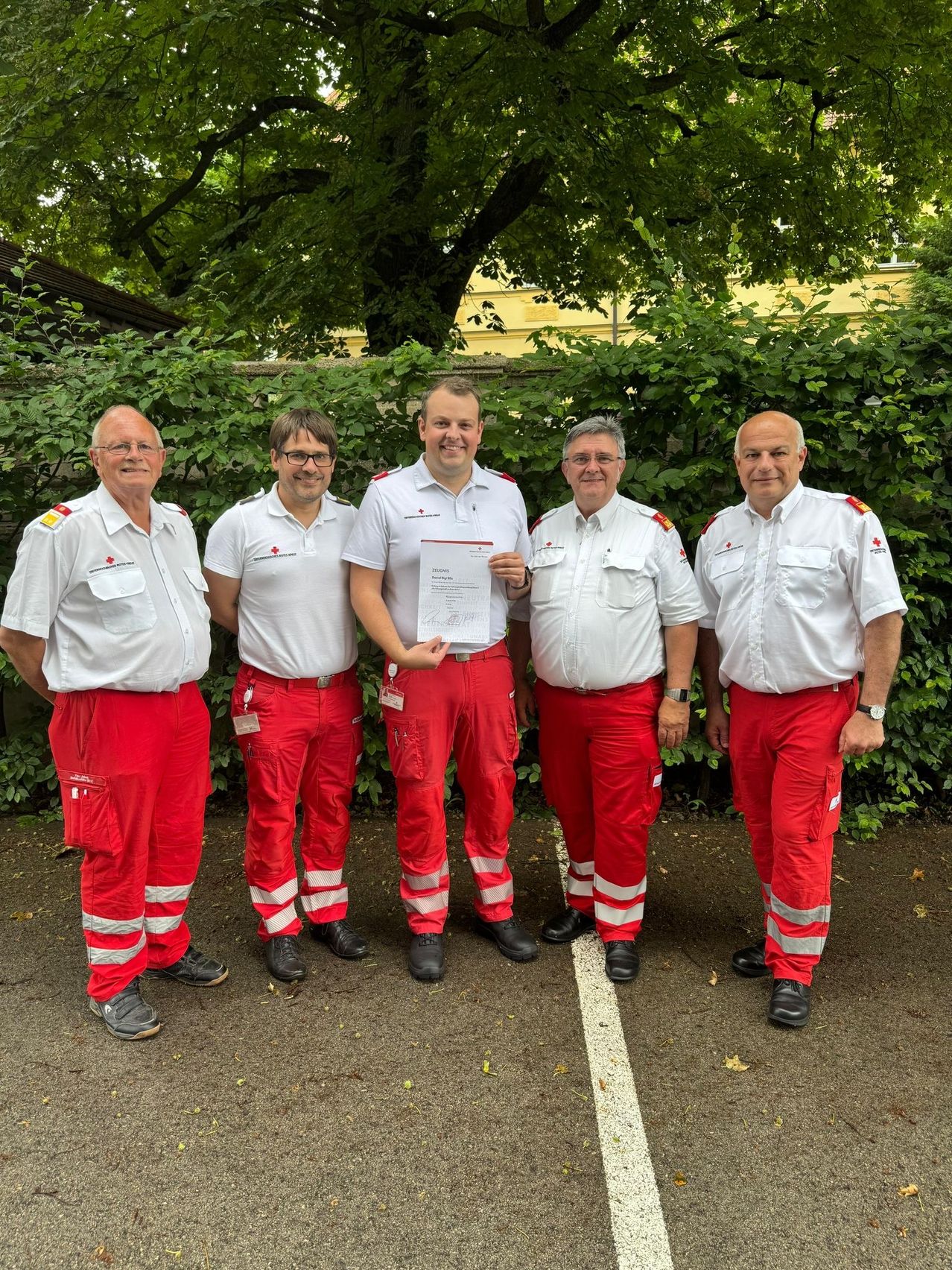 Fünf Männer in roten Hosen und weißen Hemden stehen in einer Reihe, ein Mann hält ein Zertifikat.