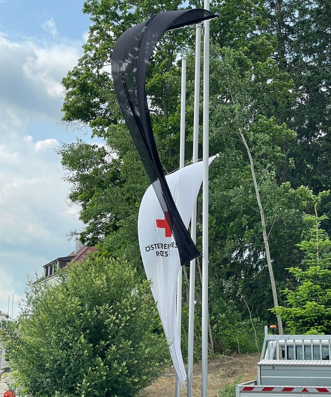 Eine schwarze und weiße Fahne mit einem roten Kreuz steht auf einem Pfosten in einem Park. Dahinter sind Bäume, ein Gebäude und ein wolkiger Himmel.