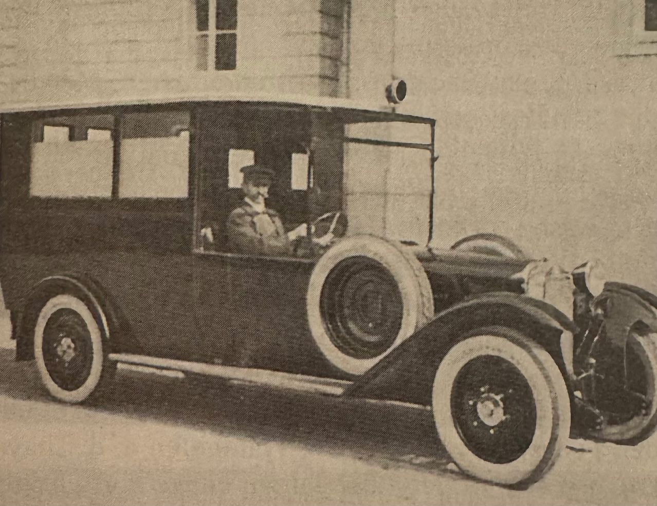 Ein schwarzer Oldtimer mit weißen Reifen steht vor einem Gebäude. Der Fahrer sitzt auf dem Fahrersitz und trägt einen Hut.