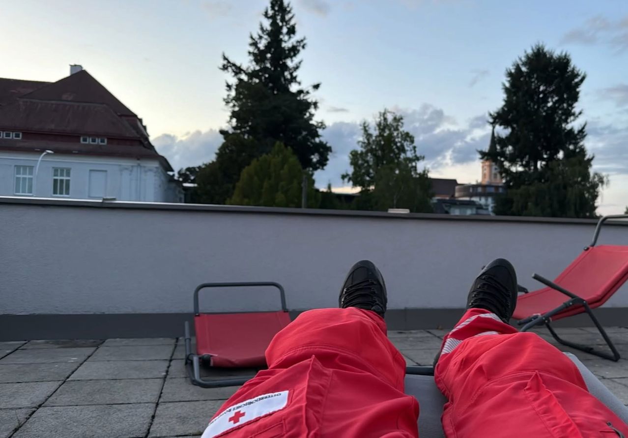 Person in roter Notfallkleidung ruht auf einem Klappstuhl auf einer Terrasse mit Bäumen und Gebäuden im Hintergrund.