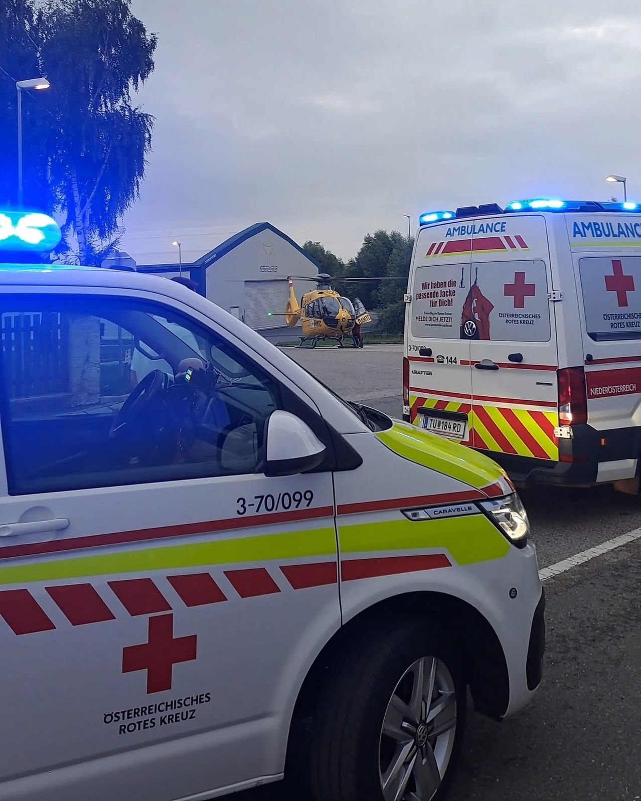 Zwei Ambulanzfahrzeuge und ein Hubschrauber sind auf einer Straße geparkt. Der vordere Krankenwagen hat ein rotes Kreuz auf der Tür. Der hintere Krankenwagen hat ein Nummernschild. Bäume und ein Gebäude sind im Hintergrund.