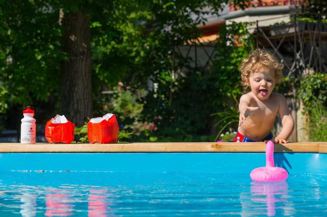 Ein junger Junge steht am Rande eines Pools, mit herausgestreckter Zunge, zwei roten Objekten in der Nähe und einem rosa Schwimmtier im Wasser.
