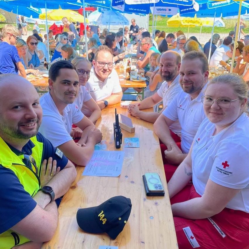 Eine Gruppe von Menschen sitzt lächelnd um einen Tisch herum, mit einer Frau in roten Hosen und einem roten Kreuz auf ihrem Shirt. Der Mann im gelben Vest schaut zur Kamera. Hinter ihnen sitzen und stehen weitere Menschen, einige mit Regenschirmen.