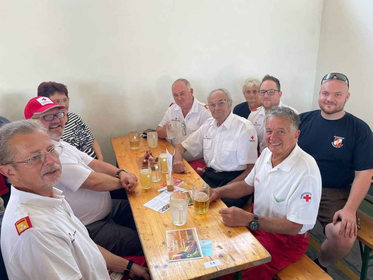 Eine Gruppe von Menschen, einschließlich Männer und Frauen, sitzt um einen holzernen Tisch mit Gläsern Bier und Essen. Sie tragen alle weiße Poloshirts mit einem roten Kreuzsymbol auf der linken Seite.