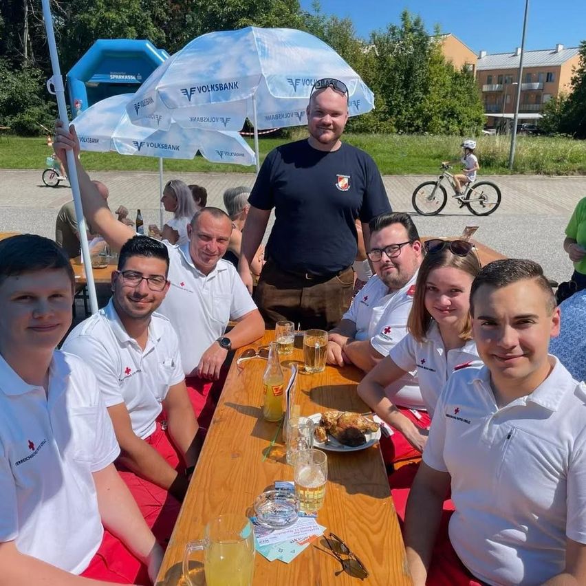 Eine Gruppe von Personen in weißen Poloshirts mit roten Shorts sitzt um einen Tisch mit Getränken und Essen. Ein Mann in einem blauen Shirt steht hinter ihnen und lächelt. Im Hintergrund fährt ein Kind mit dem Fahrrad.