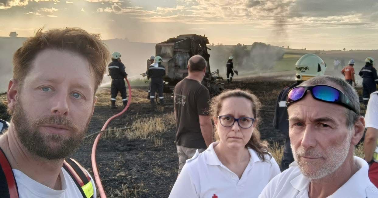 Menschen stehen auf einem Feld mit verbranntem Gras. Feuerwehrleute sprühen Wasser auf ein brennendes Fahrzeug. Der Himmel ist bewölkt.