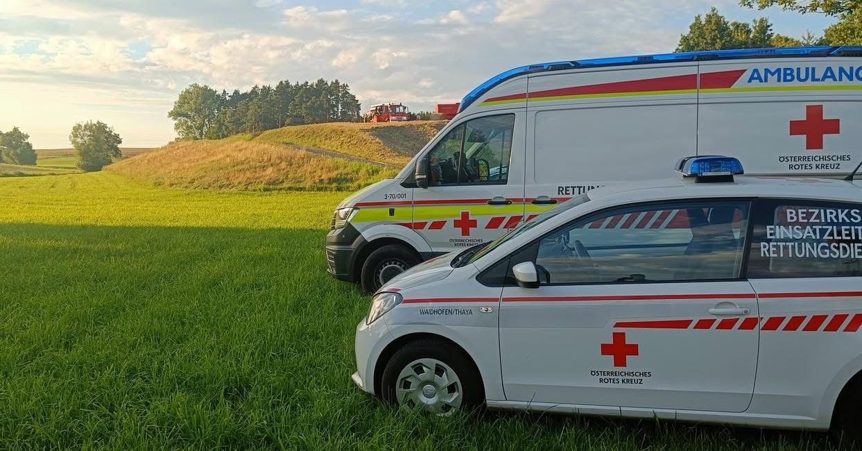 Zwei Ambulanzfahrzeuge, eines mit einem roten Kreuz an der Seite, sind auf einem Grasfeld geparkt. In der Ferne gibt es einen roten Lkw und einen Hügel mit Bäumen. Der Himmel ist teilweise bewölkt.
