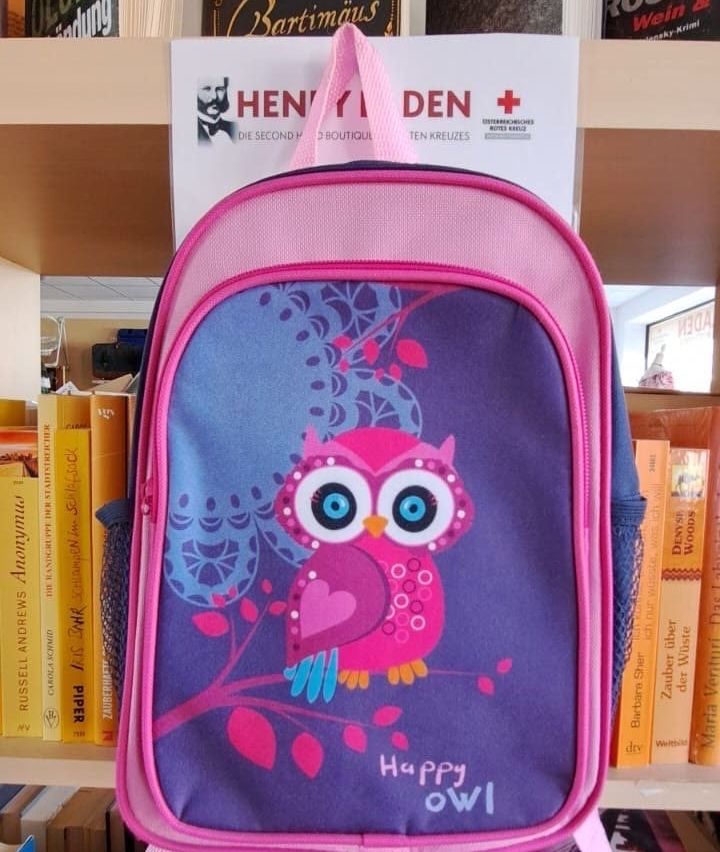 Ein rosa und lila Rucksack mit einem fröhlichen Eulendesign steht auf einem Regal zwischen anderen Büchern.