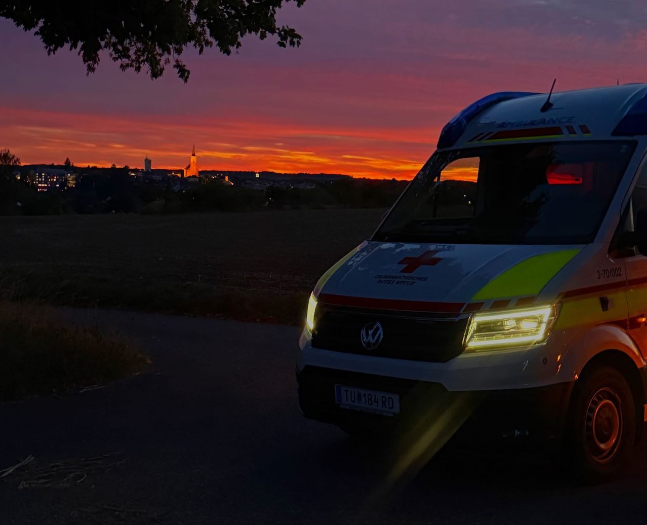 Ein Krankenwagen mit eingeschalteten Scheinwerfern steht auf einer Straße mit einer Stadtsilhouette im Hintergrund bei Sonnenuntergang.
