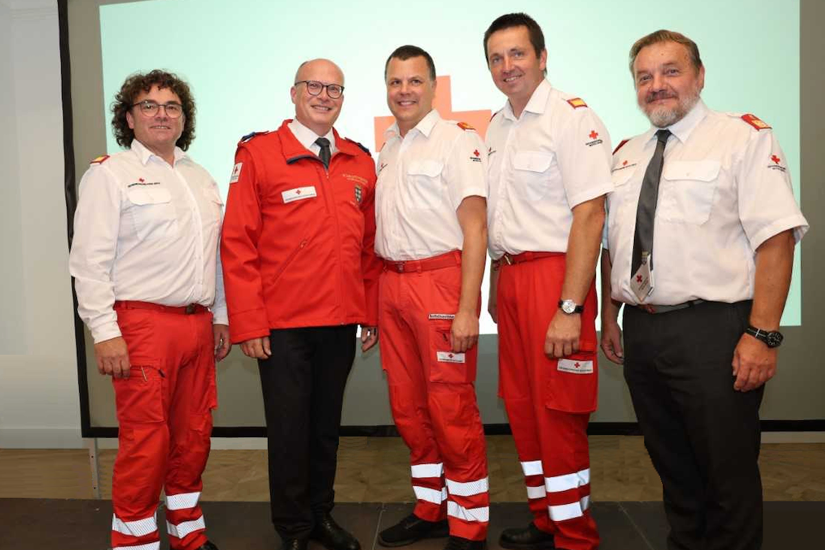 Fünf Männer in Uniformen stehen zusammen. Drei tragen rote Uniformen und zwei tragen weiße Uniformen. Sie lächeln für ein Foto.