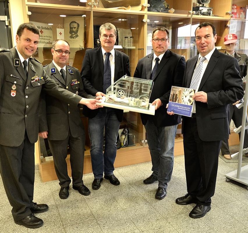 Fünf Männer in Anzügen und Krawatten, zwei in Militäruniformen, halten eine Glasbox und ein Buch. Sie posieren für ein Foto in einem Raum mit gefliestem Boden und einem Glasschrank.