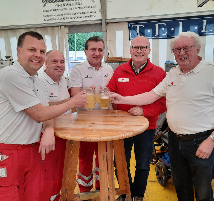 Fünf Männer in weißen und roten Uniformen lächeln und halten Gläser mit Bier in der Hand. Sie stehen um einen runden Holztisch herum. Hinter ihnen steht ein Zelt mit einem weißen und blauen Banner.