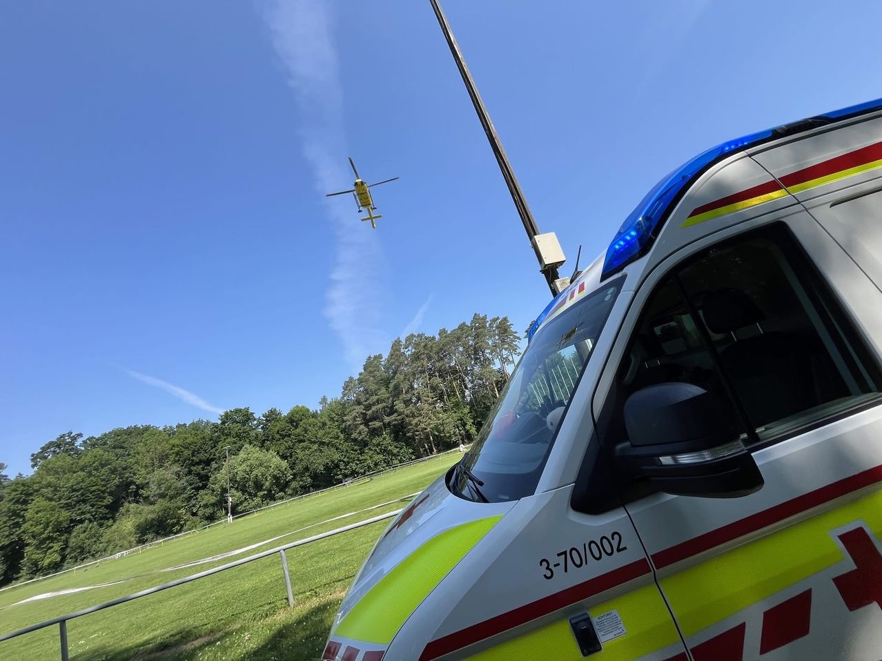 Ein Krankenwagen mit der Nummer 3-70/002 steht in der Nähe eines Feldes. Ein Hubschrauber fliegt über dem Himmel.