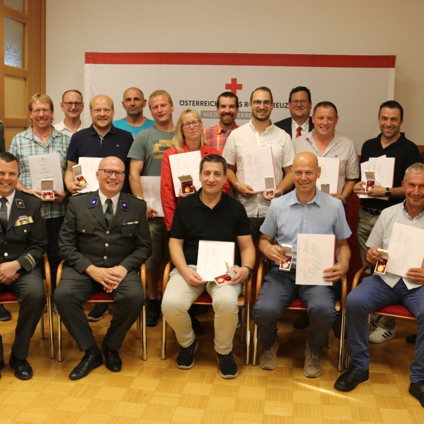 Eine Gruppe von Menschen sitzt auf Stühlen und hält Urkunden in den Händen. Ein Mann in der Mitte trägt eine Uhr. Ein Banner mit einem roten Kreuz steht hinter ihnen.
