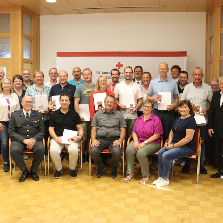 Eine Gruppe von Menschen, sowohl Männer als auch Frauen, posiert für ein Foto in einem Raum mit Holzböden. Sie tragen alle Freizeitkleidung und halten Urkunden in der Hand. Hinter ihnen befindet sich ein Banner mit einem roten Kreuz-Symbol.