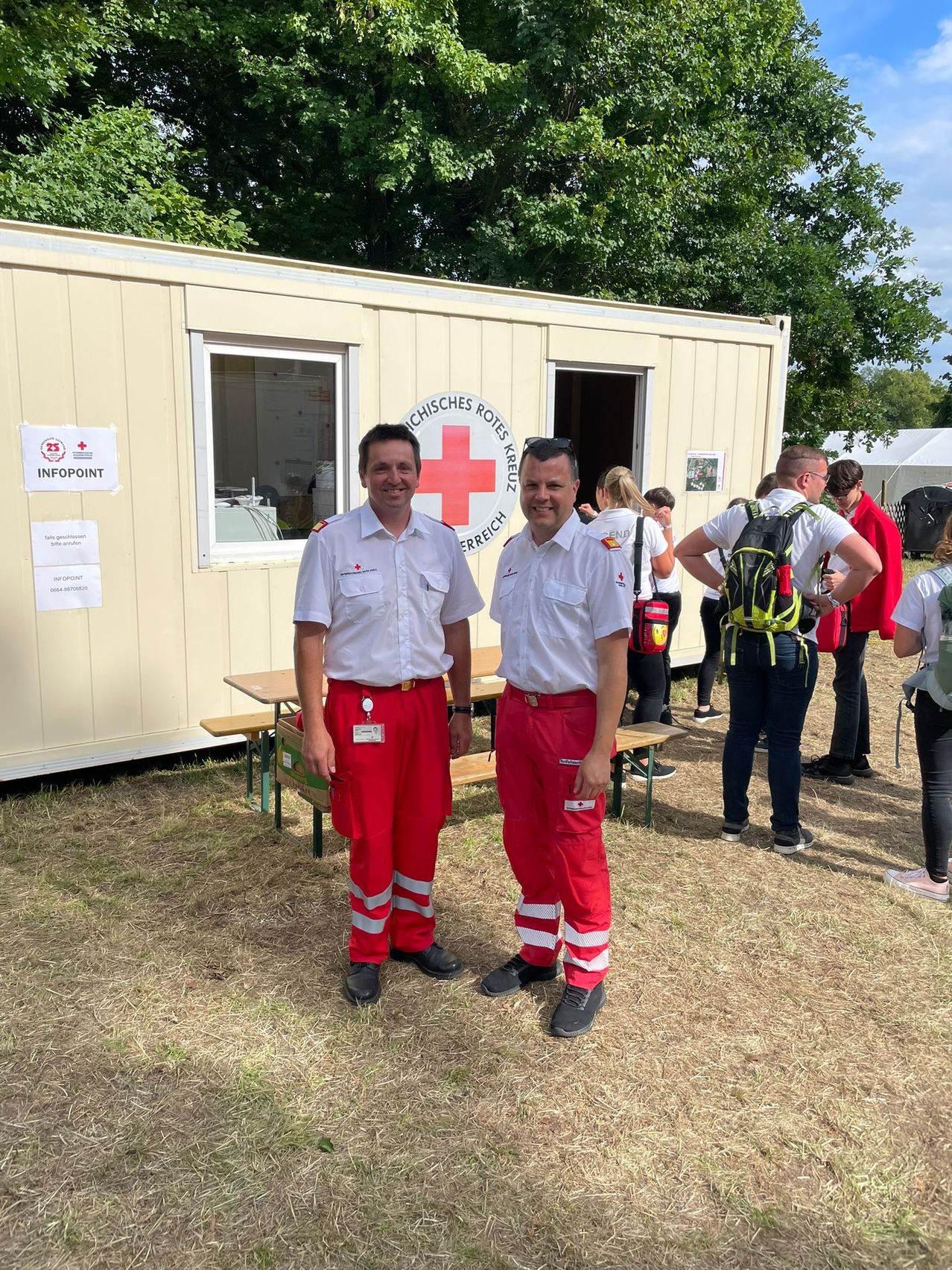 Zwei Männer stehen vor einem Gebäude mit einem roten Kreuzlogo, tragen rote Uniformen und lächeln für ein Foto. Hinter ihnen ist eine Gruppe von Menschen versammelt, einige mit Rucksäcken. Draußen steht ein Tisch mit Bänken.