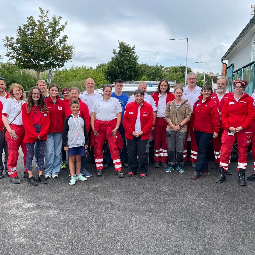 Eine Gruppe von Freiwilligen in roten Uniformen steht vor einem weißen Van. Einige tragen Brillen, und ein Junge in einem grauen Shirt steht in der Mitte. Bäume und Straßenlaternen sind im Hintergrund.