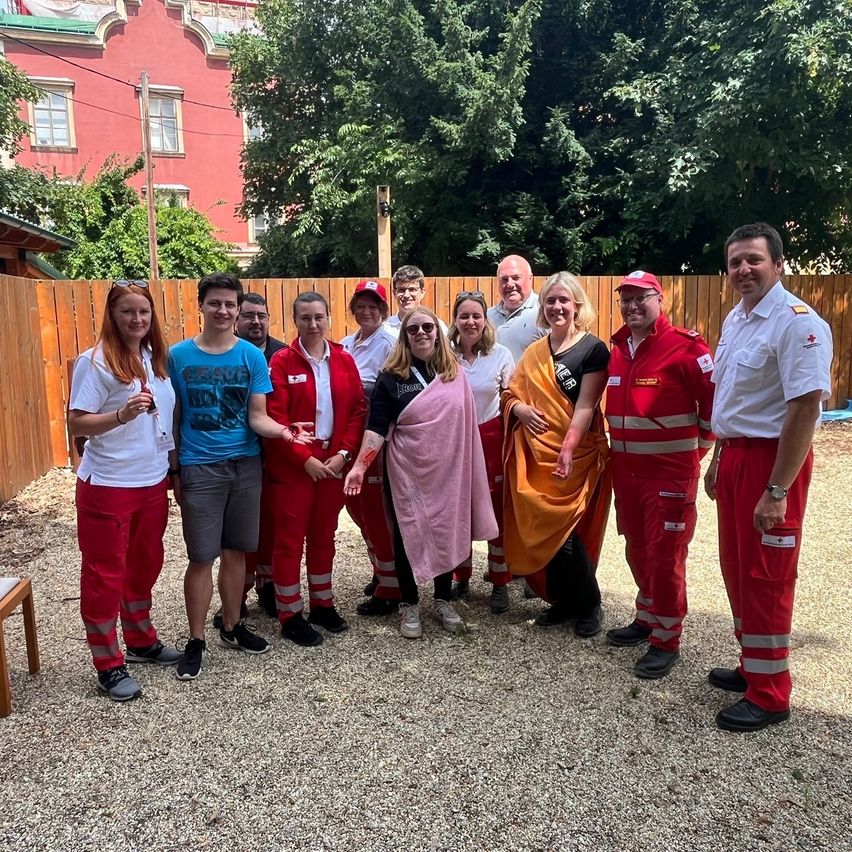 Eine Gruppe von Freiwilligen, einige in roten Uniformen, posiert für ein Foto im Freien mit einem Holzzaun und Bäumen im Hintergrund.
