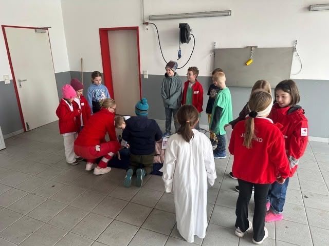 Eine Gruppe von Kindern in roten und weißen Jacken versammelt sich um eine Person auf dem Boden, wahrscheinlich übt sie Erste Hilfe in einem Raum mit Türen und einer Tafel.