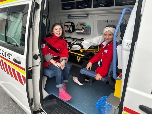 Zwei Mädchen sitzen in einem Krankenwagen, wobei eines der Mädchen einen Verband am Kopf trägt. Im Fahrzeug befinden sich medizinische Geräte.