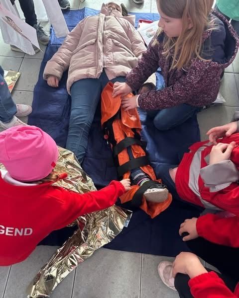 Mehrere Kinder in roten Jacken üben eine Erste-Hilfe-Prozedur an einem Dummies. Der Dummy ist in einem orangefarbenen Gurtzeug auf einem blauen Teppich befestigt.