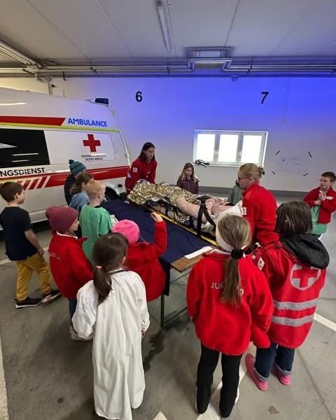Mehrere Kinder und Erwachsene beobachten eine medizinische Demonstration in einem Raum mit einem Rotkreuz-Ambulanzfahrzeug.