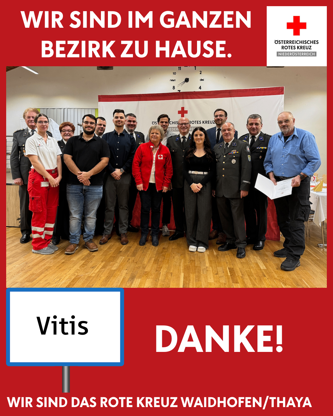 Eine Gruppe von Menschen steht vor einem Banner mit dem Roten Kreuz Logo. Zwei Personen halten Papiere. Der Text sagt 'Vitis Danke!' und 'Bezirk zu Hause.'