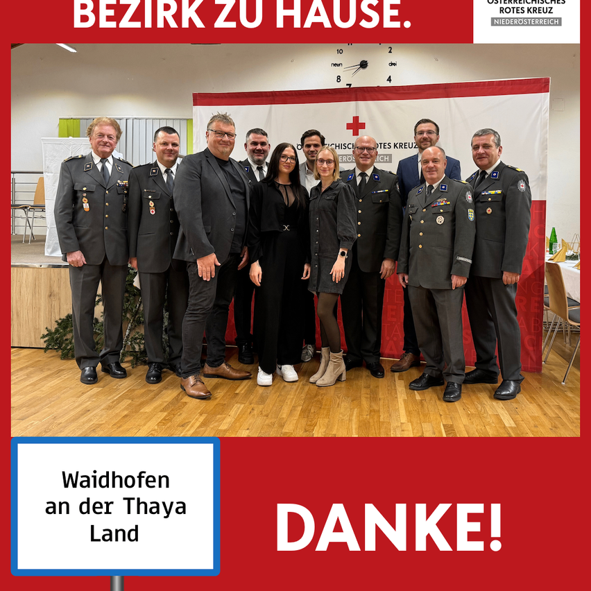 Gruppenfoto von uniformierten Personen und Zivilisten auf einer Bühne mit einem Banner, das 'Rotes Kreuz' besagt. Die Bühne ist mit Holzplanken und Grünpflanzen dekoriert. Eine Uhr und ein Schild mit der Aufschrift 'Waidhofen an der Taya Land Danke!' sind ebenfalls vorhanden.
