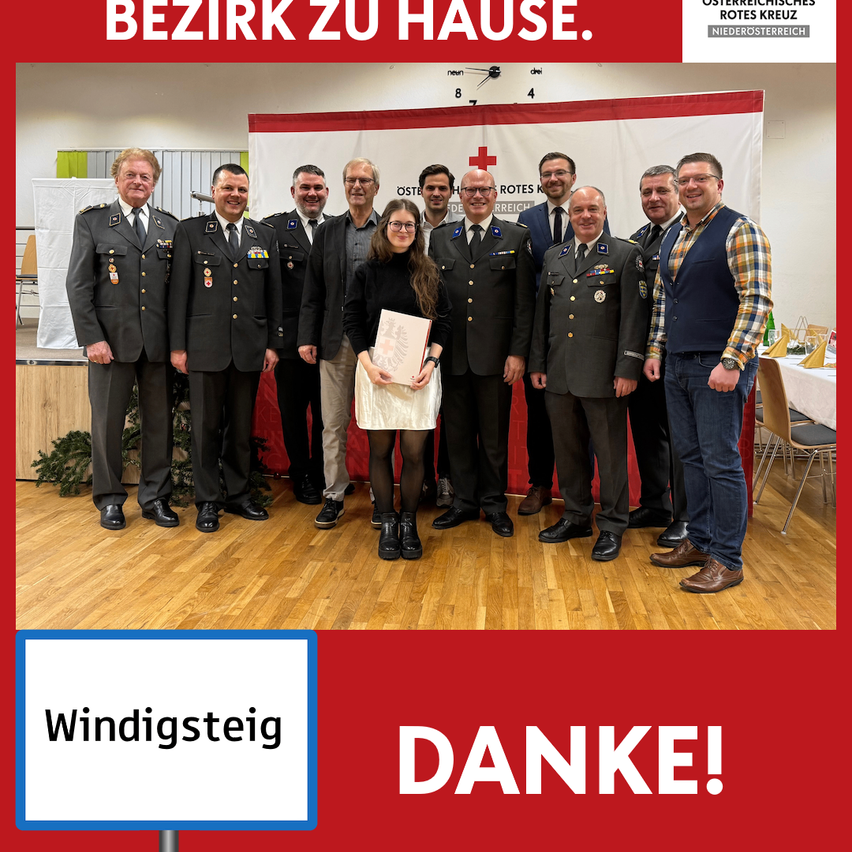Gruppenfoto von Militäroffizieren und einer Frau, die in einem Saal stehen, mit einem Banner im Hintergrund. Das Bild hat Text auf Deutsch, 'Bezirk zu Hause' und 'Windigsteig Danke!'.