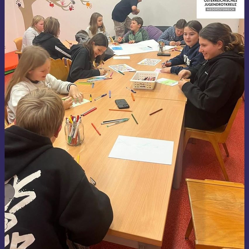 Eine Gruppe von Kindern in einem Raum ist mit einer Malaktivität beschäftigt. Sie sitzen um einen Tisch mit Papieren, Buntstiften und einem Korb. Ein Mädchen rechts lächelt und schaut ein anderes Mädchen an. Die Wand hat Zeichnungen von Händen und einem Baum.