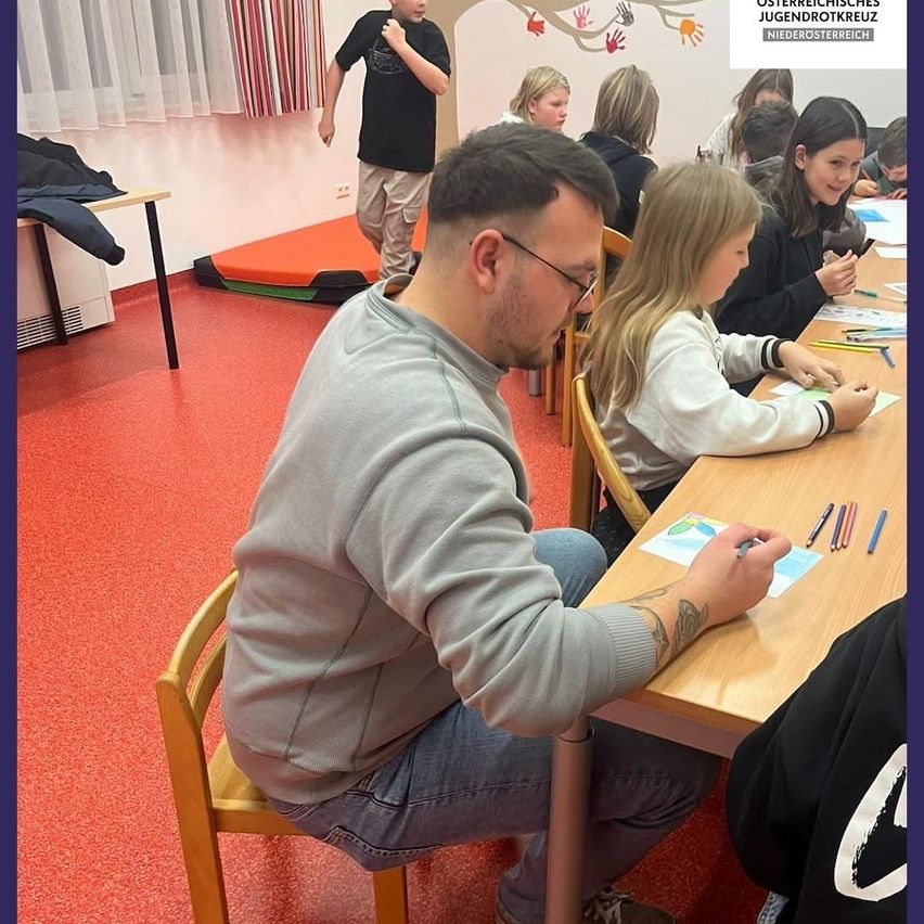 Ein Klassenzimmer mit mehreren Kindern, einem Mann, der auf Papier zeichnet, und einem Jungen, der in der Nähe der Wand läuft. Der Raum hat einen roten Boden und eine weiße Gardine.