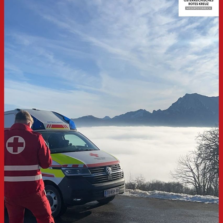 Ein Krankenwagen steht in einem nebligen Gebirgsgebiet. Der Fahrer, in einem roten Overall gekleidet, steht neben dem Fahrzeug. Die Szene spielt vor dem Hintergrund verschneiter Berge und eines blauen Himmels mit Wolken.