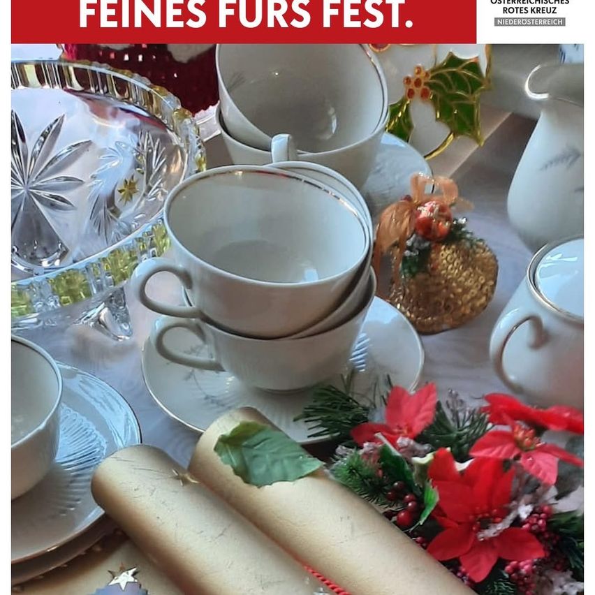 Eine festliche Tischdekoration mit weißen Tassen, Untertassen und Tellern. Der Tisch ist mit einem weißen Tuch bedeckt und mit roten Christsternen und grünen Blättern geschmückt. Es gibt eine durchsichtige Glasschale, eine Teekanne und andere weißes Geschirr. Die Worte 'Feines Furs Fest' sind auf einem roten Banner geschrieben.