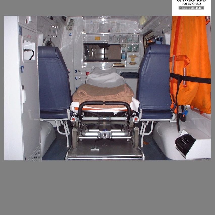 Innenraum eines Krankenwagens mit einer leeren Trage und medizinischen Geräten an den Seiten. Eine Rettungsweste hängt an der Wand.