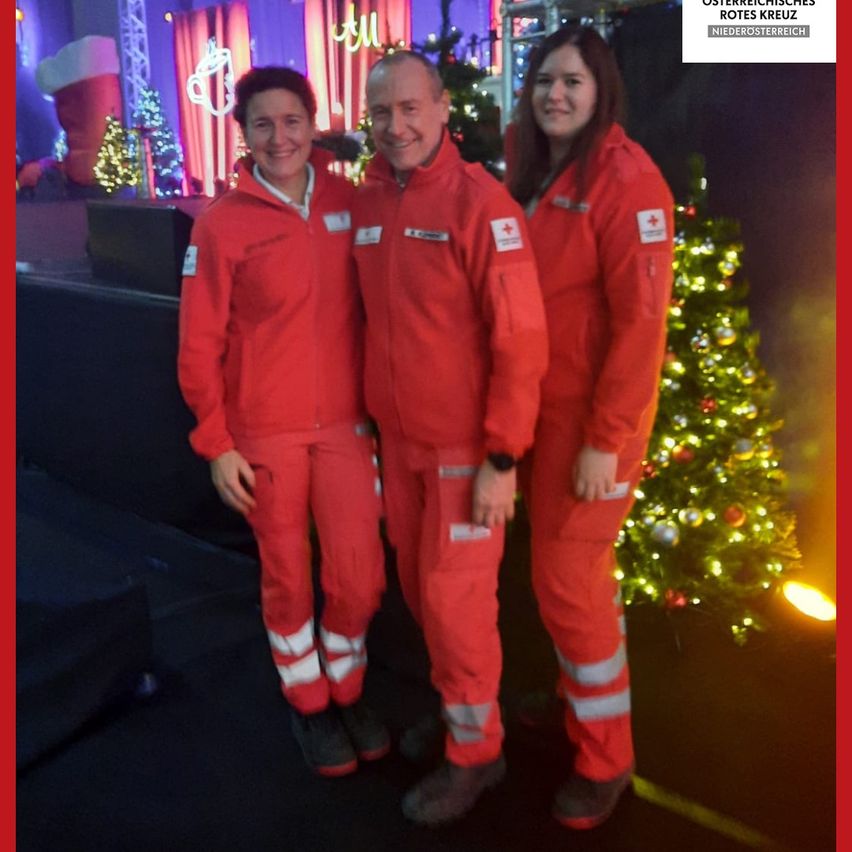 Drei Personen in roten Uniformen stehen eng nebeneinander vor einem Weihnachtsbaum. Sie scheinen für ein Foto zu posieren. Dahinter steht ein geschmückter Weihnachtsbaum und ein festlicher Hintergrund.