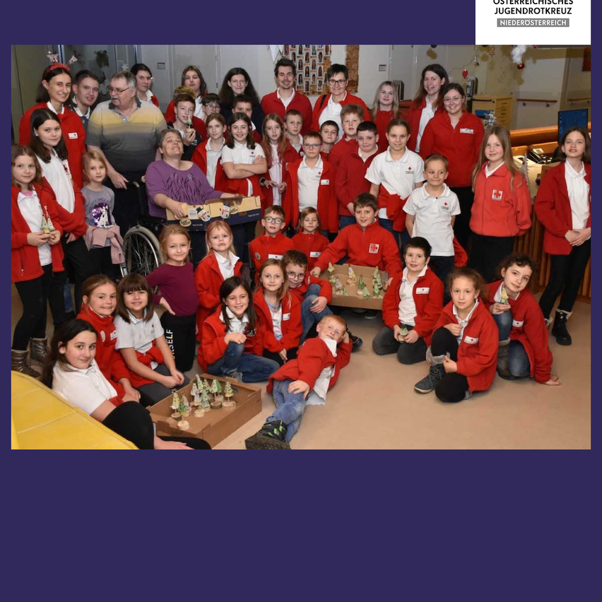 Eine Gruppe von Kindern und Erwachsenen in roten Jacken posiert für ein Foto, manche halten kleine Bäume. Hinter ihnen lächelt eine Frau im Rollstuhl. Im Hintergrund sieht man einen Raum mit weißen Wänden und einem Holzregal.