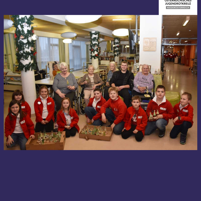 Eine Gruppe von Kindern und älteren Menschen versammelt sich in einem Raum mit Dekorationen. Sie sitzen auf dem Boden und posieren für ein Foto. Sie tragen rote Jacken. Einige von ihnen halten Geschenke. Dahinter befinden sich Säulen mit Weihnachtsdekorationen. An der Decke hängen auch Lampen. In der Ferne befindet sich eine Tür mit einer Lampe darüber.