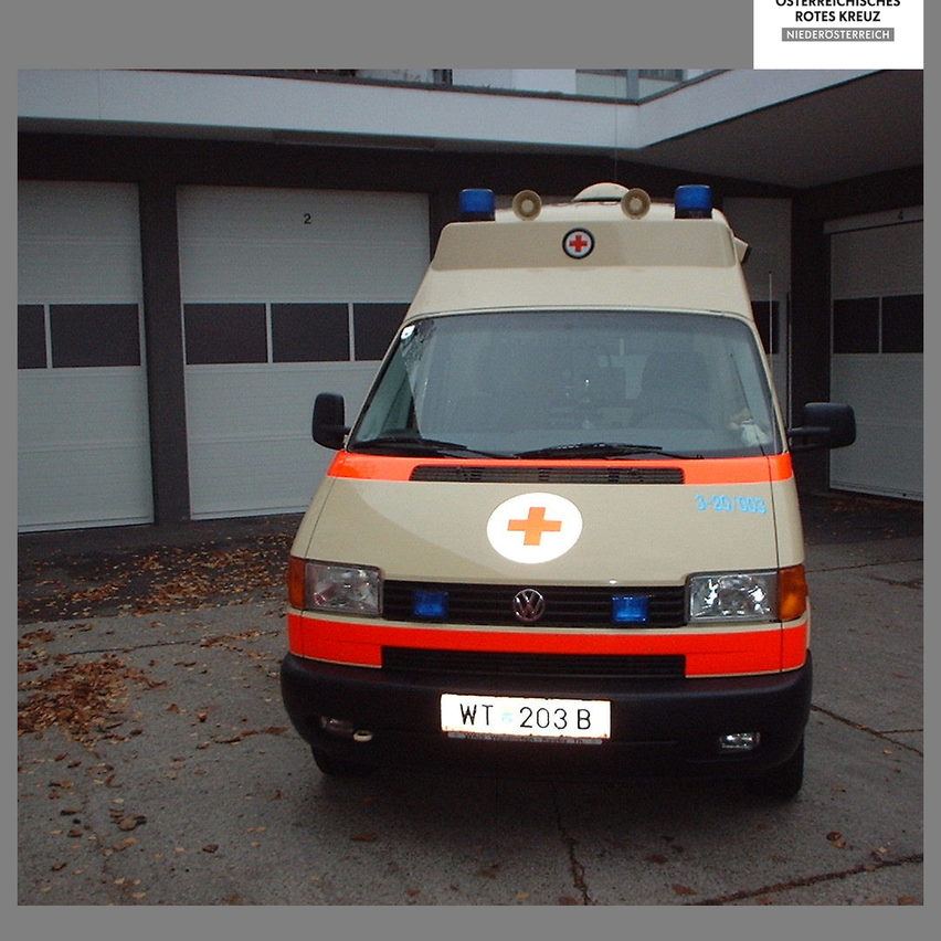Ein Krankenwagen mit einem roten Kreuzsymbol an den vorderen und seitlichen Türen steht vor einem Gebäude. Das Kennzeichen lautet 'WT 203 B'.