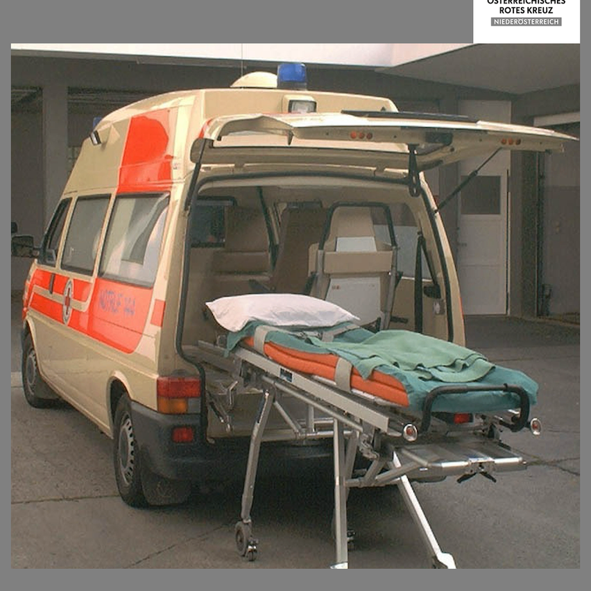 Ein Krankenwagen ist mit offener Hecktür geparkt, wobei eine leere Trage und ein Kissen im Inneren zu sehen sind. Der Krankenwagen ist weiß mit roten und orangefarbenen Streifen.