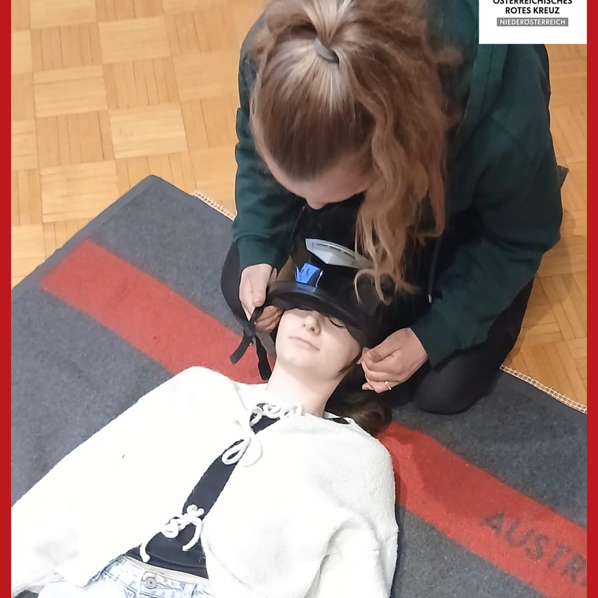 Eine Person in einer grünen Jacke justiert ein schwarzes Gerät am Kopf eines Mannequins. Das Mannequin liegt auf einem Teppich mit einem roten Streifen auf einem Holzboden.