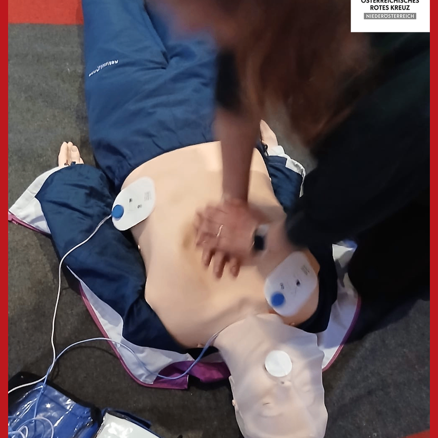 Eine Person führt Herz-Lungen-Wiederbelebung an einem Übungsdummy durch. Der Dummy liegt auf dem Boden mit einem AED-Gerät auf der Brust.