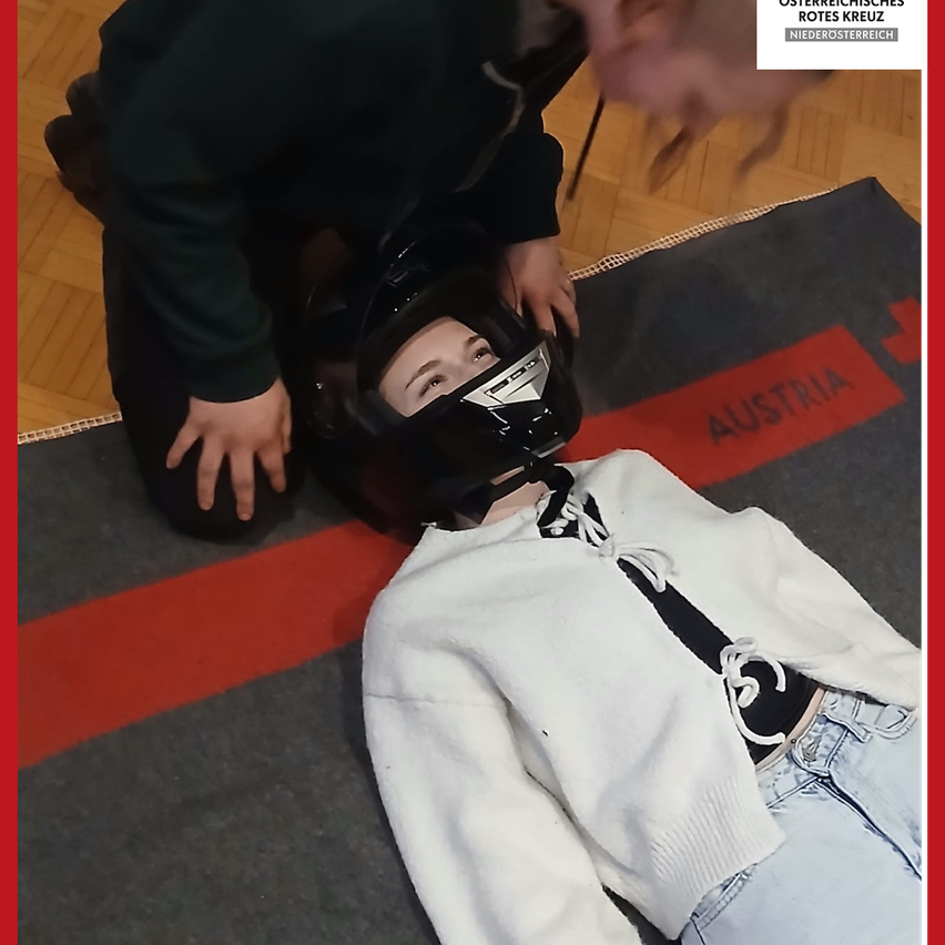 Eine Person liegt mit einem Helm auf dem Kopf auf dem Boden, eine andere Person kniet neben ihr und justiert den Helm. Sie befinden sich auf einem Teppich mit einem roten Streifen, und ein Logo mit der Aufschrift 'Austria' ist sichtbar.